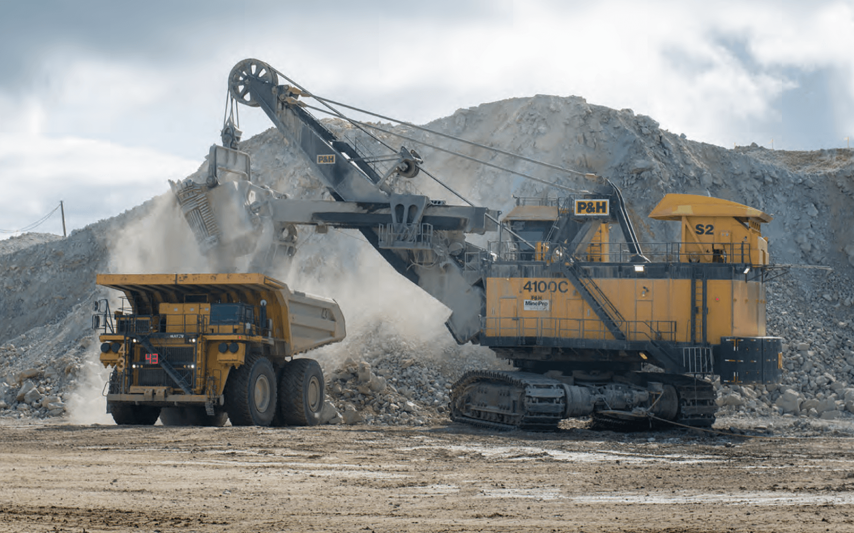 A large yellow mining truck being loaded with rock and debris by a heavy-duty excavator in an open-pit mining operation.