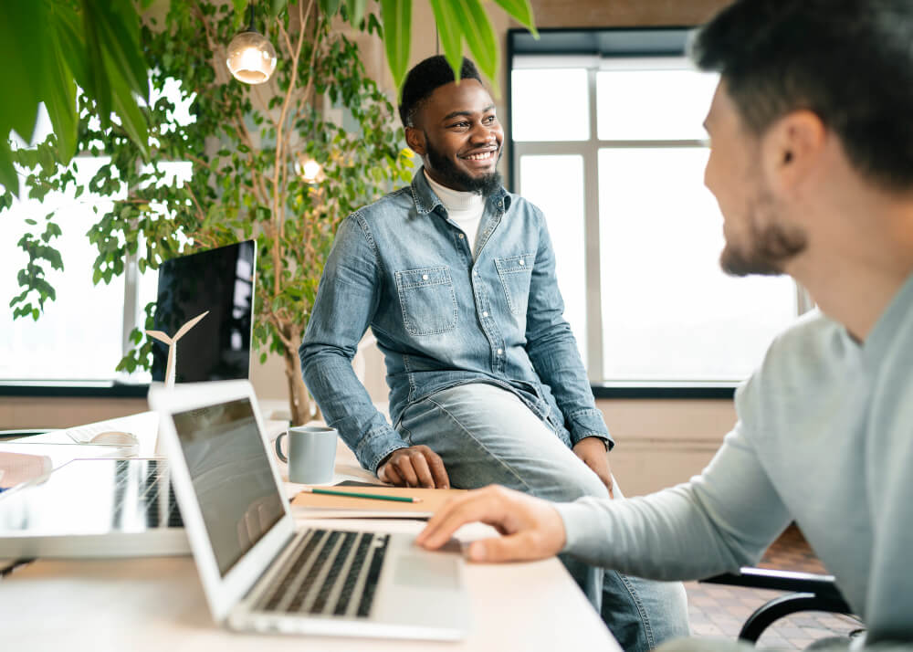 A smiling man sitting on the edge of a desk, engaged in a conversation with a colleague.